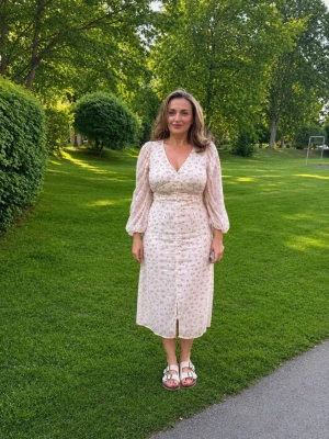 Blommig vit långklänning med knappar - Söt vit långklänning med rosa blommor och gröna detaljer. Klänningen har v-ringning, långa puffärmar och dekorativa knappar framtill. Materialet är lätt och luftigt, perfekt för varma dagar. Passformen är normal och klänningen har en slits framtill.