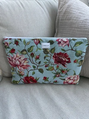 Blommig datorväska från Emelo - Snygg datorväska från Emelo i tyg med ljusblå botten och broderade blommor i rött, rosa och grönt. Väskan har en rektangulär form och stängs med dragkedja upptill. Perfekt för dig som vill ha en unik och färgglad accessoar till din laptop. Passar laptop med maxmått: 32x24cm