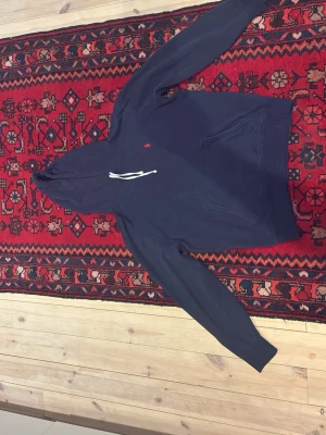 Mörkblå hoodie från Polo Ralph Lauren - Mörkblå hoodie från Polo Ralph Lauren med klassisk huva och vita dragsnören. Har en liten röd logga broderad på bröstet och en stor magficka framtill. Tillverkad i mjuk bomullsblandning som känns skön mot huden. Perfekt för en avslappnad och stilren look. Inga synliga defekter, men hör gärna av er ifall ni har några funderingar!