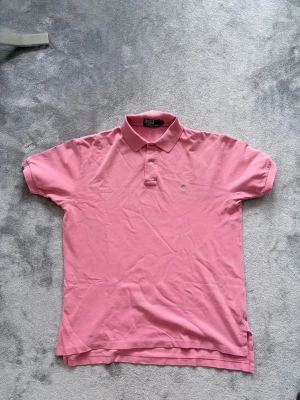 Rosa pikétröja från Polo Ralph Lauren - Klassisk rosa pikétröja från Polo Ralph Lauren med krage och korta ärmar. Tröjan har knappar framtill och en liten broderad logga på bröstet. Tillverkad i bomull och har en normal passform. Perfekt för en avslappnad och stilren look. Mått: byst 56cm, längd från axel: 66 cm