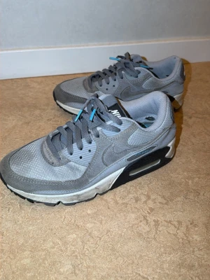 Nike Air Max 90 grå sneakers - Säljer ett par Nike Air Max 90 sneakers i olika grå nyanser med detaljer i ljusblått. Skorna har en klassisk låg siluett, mesh och mocka på ovandelen samt den ikoniska Air-sulan för extra komfort. Perfekta för dig som gillar stilrena och sportiga sneakers.