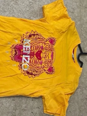 Gul Kenzo t-shirt med tigertryck - Cool gul t-shirt från Kenzo med ett stort tigertryck i rosa och rött på framsidan. T-shirten har rund hals och korta ärmar, perfekt för dig som vill sticka ut med färg och mönster. Passar dig som gillar streetstyle och grafiska prints.