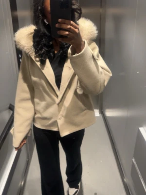 Beige kappa med fuskpäls - Stilren beige kappa med stor luva kantad av ljus fuskpäls. Modellen är rak och har knäppning framtill. Perfekt för dig som vill ha en klassisk men trendig look. Passar till många olika outfits och håller dig varm under kyliga dagar.
