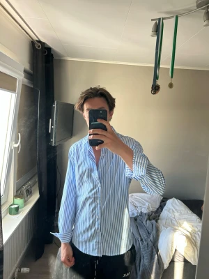 Blåvit randig skjorta med knappar - Säljer en stilren blåvit randig skjorta med klassisk krage och knäppning framtill. Skjortan har lång ärm och en relaxed passform som ger en chill vibe. Perfekt att matcha med jeans eller chinos för en clean look. Väldigt snygg och fräsch och helt utmärkt att ha på sommaren med kompisarna. Väldigt skön och som sagt väldigt snygg 