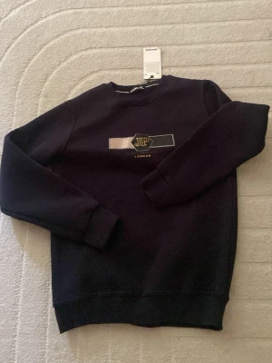 Svart sweatshirt från Jourmate JP London - Svart sweatshirt från Jourmate med JP London-logga i guld och vitt framtill. Tröjan har rund halsringning, långa ärmar och ribbade muddar. Perfekt för en stilren och avslappnad look.