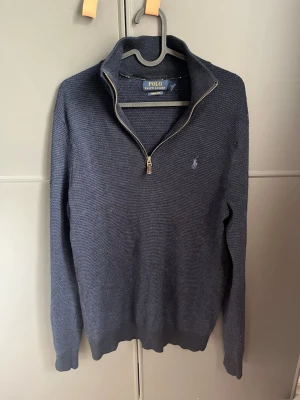 Mörkblå stickad tröja Polo Ralph Lauren - Mörkblå stickad tröja från Polo Ralph Lauren med halv dragkedja och klassisk broderad logga på bröstet. Tröjan har ribbade muddar och krage, perfekt för lager på lager. Materialet är mjukt och strukturerat för en avslappnad men snygg look. De finns ett litet hål vid axeln, kan gå ner i pris vid snabb affär.