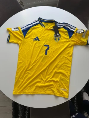 Sverige matchtröja Adidas gul/blå - Säljer en officiell gul matchtröja från Sveriges landslag med blå detaljer, nummer 7 och namnet Bergvall på ryggen. Tröjan har korta ärmar, broderad landslagssköld och Adidas-logga. Tillverkad i lätt funktionsmaterial, perfekt för fotboll eller träning.