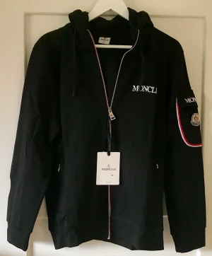 Svart Moncler hoodie med dragkedja+ byxor - Säljer en svart hoodie från Moncler med dragkedja framtill, huva med dragsko och vita Moncler-loggor på bröstet och ärmen. Matchande byxor medföljer.Detaljer i rött, vitt och blått längs dragkedjan och på ärmen. Perfekt för dig som gillar exklusiva streetwear-plagg.