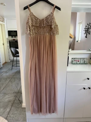 Lång champagnefärgad klänning med paljetter - Elegant långklänning i champagnefärg med smala axelband och glittrig paljettöverdel. Kjolen är i mjukt, tunt tyg som faller snyggt och ger en romantisk look. Perfekt för bal eller festliga tillfällen.
