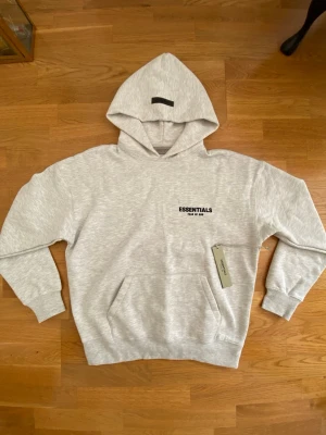 Grå Essentials Fear of God hoodie - Snygg ljusgrå hoodie från Essentials Fear of God med svart logga på bröstet och stor text på ryggen. Klassisk känguruficka framtill, ribbade muddar och stor huva. Perfekt för en avslappnad streetwear-look.