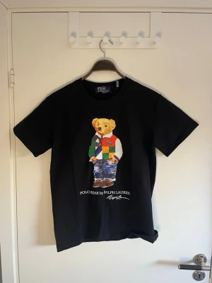 Svart Polo Bear t-shirt från Ralph Lauren - Säljer en svart t-shirt från Ralph Lauren med den ikoniska Polo Bear-trycket på bröstet. Björnen har färgglada kläder och trycket är detaljerat. T-shirten har rund hals och korta ärmar. Perfekt för dig som vill ha en klassisk men lekfull look.