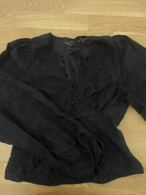 Svart blus med knappar och puffärm - Stilren svart blus från Gina med blommönster i tyget. Blusen har långa puffärmar, v-ringning och knappar framtill. Materialet är tunt och lätt transparent. 