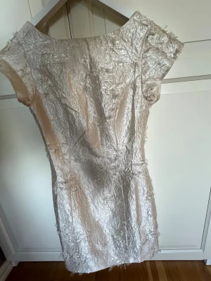 Champagnefärgad kort klänning - Säljer en champagnefärgad kort klänning med unik textur och lätt glans. Klänningen har korta ärmar och en figurnära passform. Materialet känns exklusivt och skimrande, perfekt för dig som vill sticka ut.