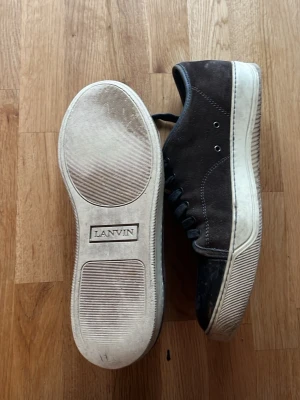 Bruna Lanvin sneakers i mocka - Snygga bruna sneakers från Lanvin med ovandel i mocka och tåparti i svart läder. Skorna har svarta skosnören, metallöljetter och vit sula med Lanvin-logga undertill. Perfekta för dig som gillar stilrena och exklusiva sneakers.