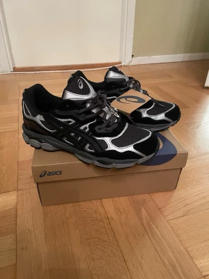 Svarta och silver Asics sneakers - Snygga Asics sneakers i svart och silver med meshpaneler och detaljer i syntetmaterial. Skorna har en chunky sula och klassisk Asics-logga på sidan. Perfekta för dig som gillar sportig och trendig stil.
