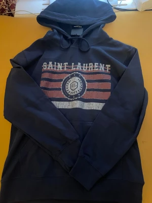 Mörkblå Saint Laurent hoodie - Snygg mörkblå hoodie från Saint Laurent med tryck framtill i vitt och rött. Hoodien har en klassisk huva med dragsko och en stor magficka. Materialet känns mjukt och bekvämt, perfekt för en avslappnad stil. Trycket har en vintage-look med text och emblem.