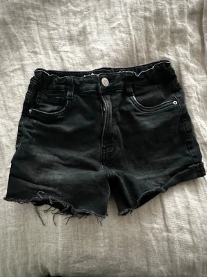 Svarta denimshorts med rå kant - Snygga svarta denimshorts med hög midja och rå, fransig kant nertill. Klassisk femficksmodell med knapp och dragkedja framtill. Perfekta för en avslappnad och trendig sommarlook. Strl 158.