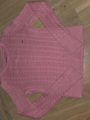 Rosa kabelstickad tröja Tommy Hilfiger - Säljer en rosa kabelstickad tröja från Tommy Hilfiger med rund halsringning och diskret logga broderad på bröstet. Tröjan har långa ärmar och ribbade muddar vid ärmslut och nederkant. Perfekt för dig som gillar klassisk och stilren design.