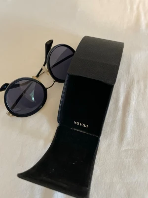 Runda svarta solglasögon från Prada - Snygga runda solglasögon från Prada med svarta bågar och mörka glas. Metallbågarna har en elegant och stilren design som ger en cool vibe. Kommer med originalfodral från Prada. Perfekta för dig som vill ha en trendig accessoar.