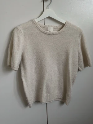 Beige stickad t-shirt från H&M - Mjuk och stilren beige stickad t-shirt från H&M med korta ärmar och rund halsringning. Tröjan har ribbade kanter vid hals, ärmslut och nederkant. Perfekt för lager på lager och enkel att matcha med olika outfits.