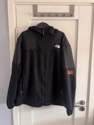 Svart hoodie-cardigan från The North Face - Snygg svart hoodie-cardigan från The North Face med dragkedja framtill och huva. Jackan har röda detaljer på dragkedjorna och logga på bröstet samt ärmen. Materialet är en mix av slät och mesh-liknande textur, perfekt för lager-på-lager och streetwear.