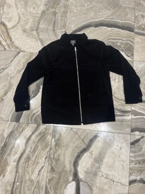Svart manchester overshirt från H&M - Säljer en svart overshirt från H&M i manchester med regular fit. Jackan har två bröstfickor, klassisk krage och hel dragkedja framtill. Perfekt att slänga över en hoodie eller t-shirt för en chill look.