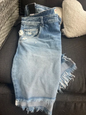 Blå jeansshorts med fransig kant - Säljer jeanskjol från Replay med slitna detaljer och fransig kant nedtill. Klassisk femficksmodell och knappgylf. Perfekta för dig som gillar en avslappnad och trendig stil.