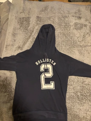 Svart Hollister hoodie med tryck - Mörkblå hoodie från Hollister med stor vit siffra 2 och texten 'Hollister' på bröstet. Klassisk huva med dragsko och långa ärmar. Perfekt för en avslappnad och sportig stil. Materialet känns mjukt och bekvämt, perfekt för vardagsbruk.