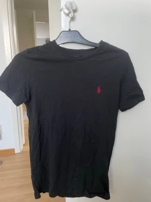 Svart t-shirt från Ralph Lauren - Klassisk svart t-shirt från Ralph Lauren med korta ärmar och rund hals. På bröstet finns den ikoniska röda Polo-loggan broderad. Tillverkad i mjuk bomull som känns skön mot huden. Perfekt basplagg för en clean och stilren look. Ursprungligen är den designad för män men jag har också använt den och den har suttit bra!