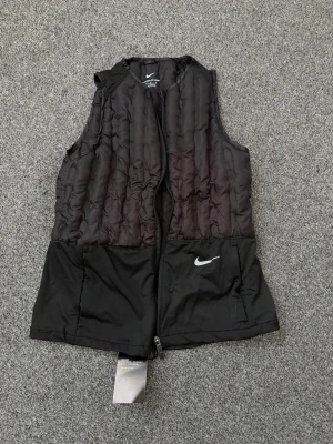 Svart dunväst från Nike Therma-FIT - Snygg svart dunväst från Nike med Therma-FIT-teknologi. Västen har quiltad ovandel och slät nederdel, dragkedja framtill och en vit Nike-logga på bröstet. Perfekt för lager på lager och sportiga outfits.