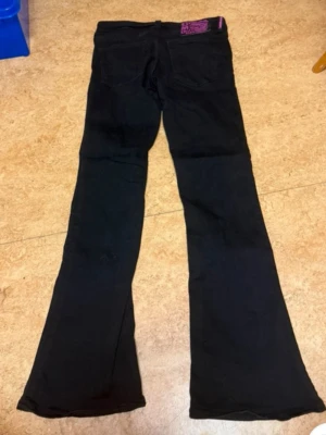 Svarta utsvängda jeans Cheap Monday - Säljer ett par svarta jeans från Cheap Monday med utsvängda ben och klassisk femficksmodell. Jeansen har en lila patch med logga bak i midjan och är tillverkade i ett stretchigt bomullsmaterial. Perfekta för dig som gillar retrostil och vill sticka ut.
