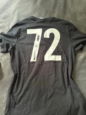 Svart Nike t-shirt med tryck - Snygg svart t-shirt från Nike med stort vitt tryck av siffran 72 på ryggen och Nike-logga samt text på bröstet. Klassisk rund hals och korta ärmar. Perfekt för dig som gillar sportig och stilren design.