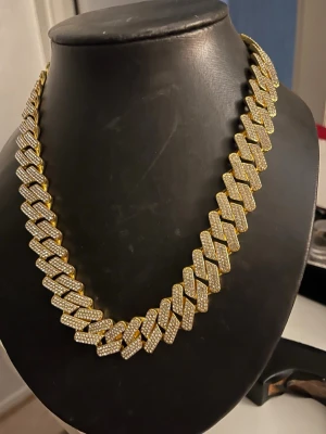 Guldfärgat cuban chain halsband - Snyggt guldfärgat halsband i cuban chain-stil med stora länkar täckta av glittrande stenar som ger en lyxig känsla. Perfekt för dig som vill sticka ut och addera lite extra bling till din outfit. Halsbandet har en chunky design och känns riktigt trendigt.