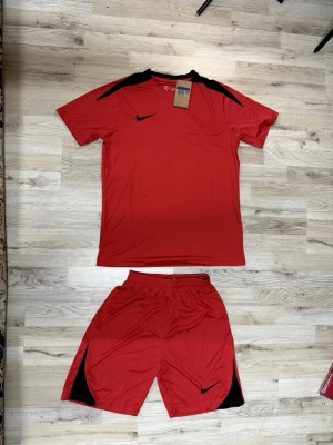 Rött Nike träningsset herr - Säljer ett rött träningsset från Nike med t-shirt och shorts. Setet har svarta detaljer på axlar och sidor samt Nike-logga på både tröja och shorts. Perfekt för fotboll eller gym. Materialet är lätt och andas, vilket gör det bekvämt under träning.
