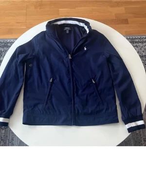 Marinblå vindjacka från Polo Ralph Lauren - Snygg marinblå vindjacka från Polo Ralph Lauren med vit rand på krage och ärmslut. Jackan har dragkedja framtill, två snedställda fickor med dragkedja och en liten logga på bröstet. Perfekt för blåsiga dagar och sportig stil. Den är i bra skick men om något så kan det vara en liten missfärgning på det vita i nacken.