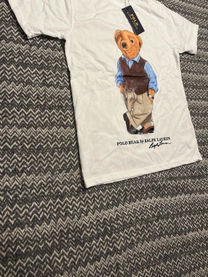 Vit Polo Bear t-shirt från Ralph Lauren - Säljer en vit t-shirt från Ralph Lauren med den ikoniska Polo Bear-trycket framtill. T-shirten har rund hals och korta ärmar. Perfekt för dig som gillar klassisk och stilren med en kol design.den är oanvänd och i storlek s