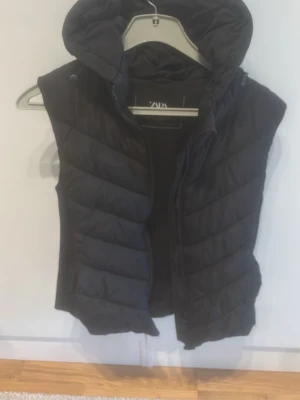 Svart dunväst från Zara med huva - Snygg svart dunväst från Zara med quiltad design och avtagbar huva. Västen har dragkedja framtill och en modern, något figurnära passform. Perfekt för lager-på-lager och enkel att matcha med olika outfits.