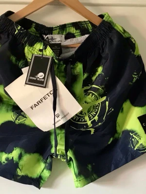 Stone Island badbyxor neon och svart - Säljer ett par badbyxor från Stone Island i en riktigt fet färgkombination av neon-grönt och svart med coolt batikmönster och stora loggor. Resår i midjan och snörning framtill. Materialet är snabbtorkande polyester och har meshfoder inuti.