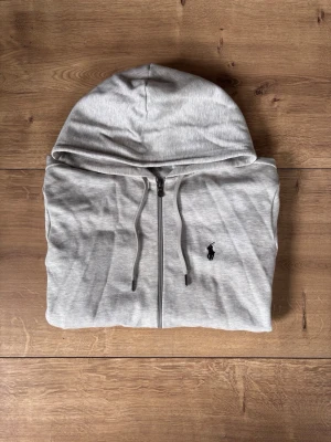 Grå hoodie från Polo Ralph Lauren - Snygg grå zip-hoodie från Polo Ralph Lauren med klassisk broderad logga på bröstet. Hoodien har dragkedja, dragsko i huvan och är gjord i mjuk bomullsblandning. Perfekt för en chill och stilren look.
