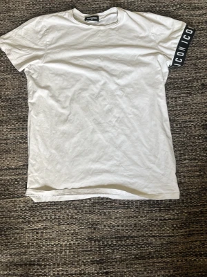 Vit t-shirt från Dsquared2 - Säljer en vit t-shirt från Dsquared2 med trycket 'ICON' på ena ärmen. Klassisk rund hals och korta ärmar. T-shirten är i mjuk bomull och har en clean look med en cool detalj på ärmen.