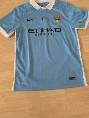 Manchester City matchtröja Nike - Ljusblå Manchester City matchtröja från Nike med vit krage och vita detaljer på ärmarna. Tröjan har klubbmärke och sponsortryck på bröstet samt Dri-FIT-material som andas. Perfekt för fotbollsträning eller att bära på läktaren.