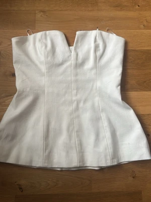 Vit axelbandslös topp från Zara - Säljer en stilren vit axelbandslös topp från Zara i storlek XS. Toppen har en snygg, figurnära passform och en lätt struktur i tyget. Perfekt att matcha med både jeans och kjol för en trendig look.