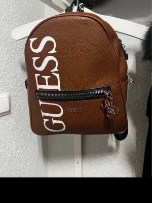 Brun ryggsäck från Guess - Snygg brun ryggsäck från Guess i konstläder med stort vitt logotryck på framsidan. Väskan har en framficka med dragkedja, metallberlocker och justerbara axelband med Guess-tryck. Perfekt storlek för det viktigaste och ger en trendig look.