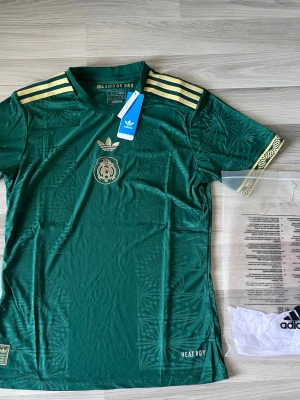 Grön Mexiko tröja från Adidas - Snygg grön Mexiko fotbollströja från Adidas med guldfärgade detaljer och diskret mönster i tyget. Tröjan har korta ärmar, rund hals och tre guldiga ränder på axlarna. Mexikos emblem i guld på bröstet och 'HEAT.RDY' tryckt nedtill.