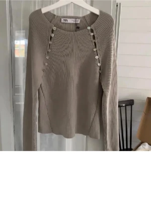 Beige stickad tröja från Zara - Snygg beige stickad tröja från Zara med dekorativa knappar längs axlarna och lång ärm. Tröjan har en rak passform och ribbstickad struktur som ger en modern look. Perfekt för dig som gillar stilrena och trendiga plagg.