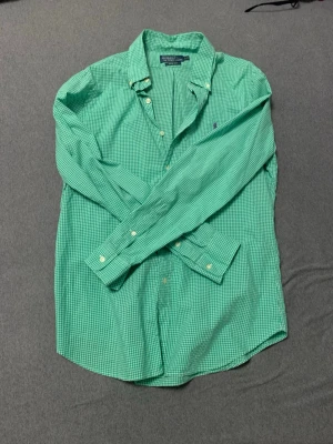 Grön rutig skjorta från Polo Ralph Lauren - Snygg grön rutig skjorta från Polo Ralph Lauren med klassisk krage och lila broderad logga på bröstet. Skjortan har lång ärm, knappar framtill och är tillverkad i bomull. Perfekt för dig som gillar stilrena och tidlösa plagg.