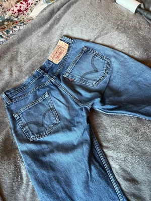 Levi's blå raka jeans med slitningar - Säljer ett par klassiska blå Levi's jeans med perfekta passformen, raka ben och slitna detaljer på framsidan. Jeansen har två bakfickor med den ikoniska Levi's-sömmen och en röd etikett. Tillverkade i denim, perfekt för en avslappnad och trendig look.