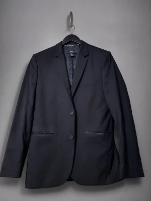 Svart kavaj från H&M Regular Fit - Stilren svart kavaj från H&M i modellen Regular Fit. Klassisk design med två knappar framtill, smala slag och tre fickor. Perfekt för en dressad look och enkel att matcha med skjorta eller t-shirt. Tillverkad i ett slätt tyg som ger en snygg siluett.