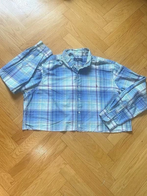 Blå rutig croppad skjorta Polo Ralph Lauren - Säljer en croppad skjorta från Polo Ralph Lauren i blått och vitt rutigt mönster. Skjortan har långa ärmar, klassisk krage och knappar framtill. Perfekt för en avslappnad och trendig stil.