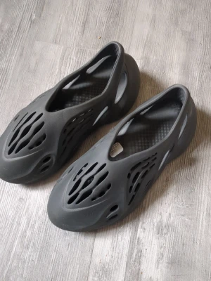 Svarta foam tofflor med hålmönster - Säljer ett par svarta tofflor i foam-material med futuristiskt hålmönster och rundad form. Lätta och bekväma att bära, perfekta för chill dagar hemma eller ute. Snygg design som sticker ut och passar till streetwear-stil har gått på ett häftstift med en av dom, synns knappt och påverkar inte Användningen.